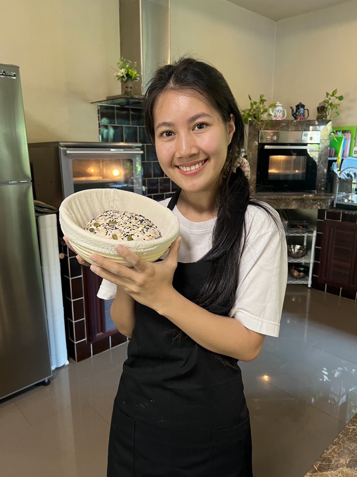 A smiling baker holding a cane proofing basket with a seeded sourdough
